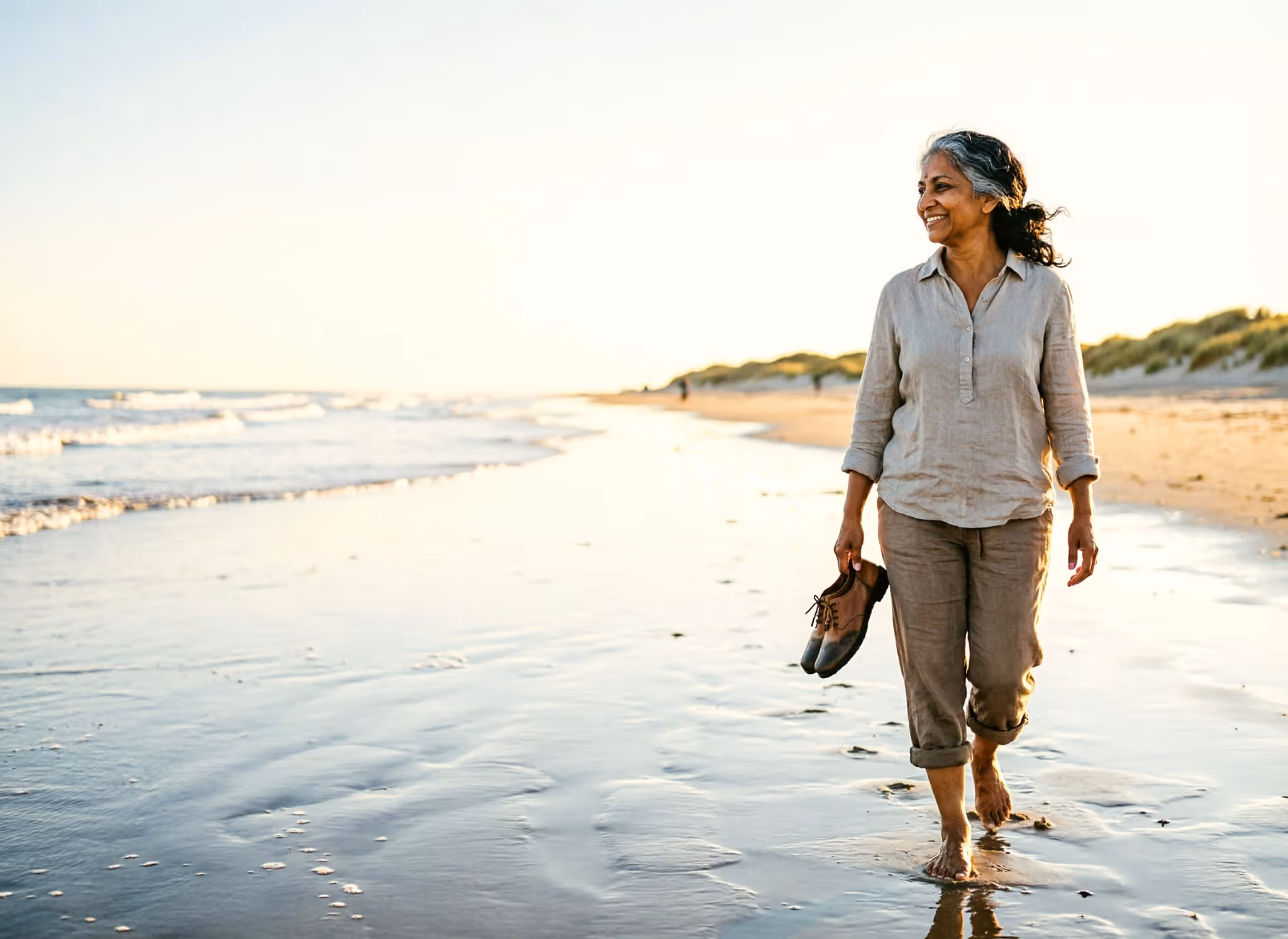 A woman in her fifties walks along the North Sea shoreline with her shoes in one hand