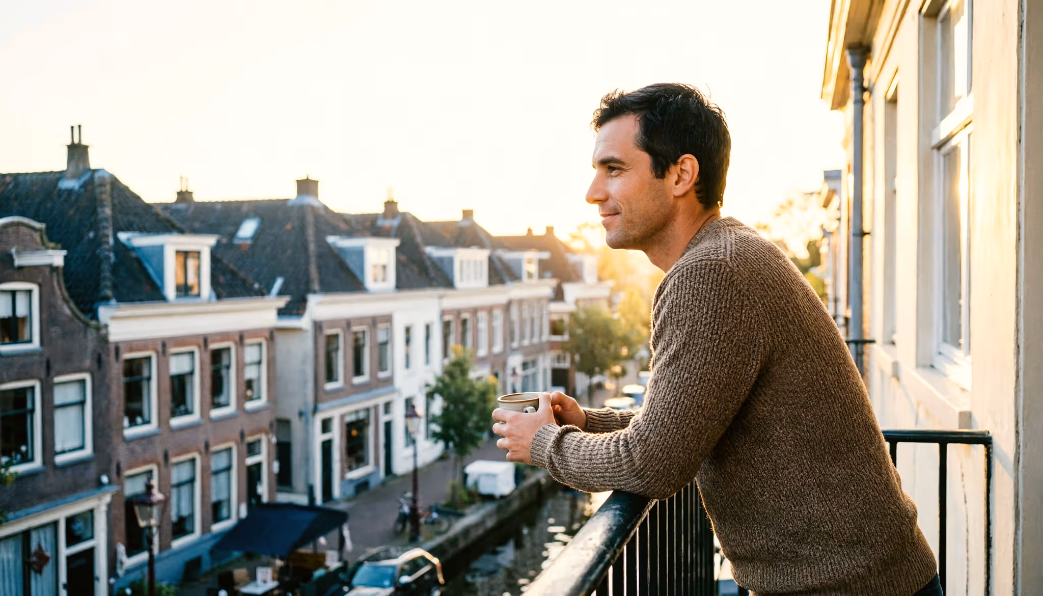 Een man ontspant met een kop koffie op zijn balkon, uitzicht over Nederlandse daken in het ochtendlicht