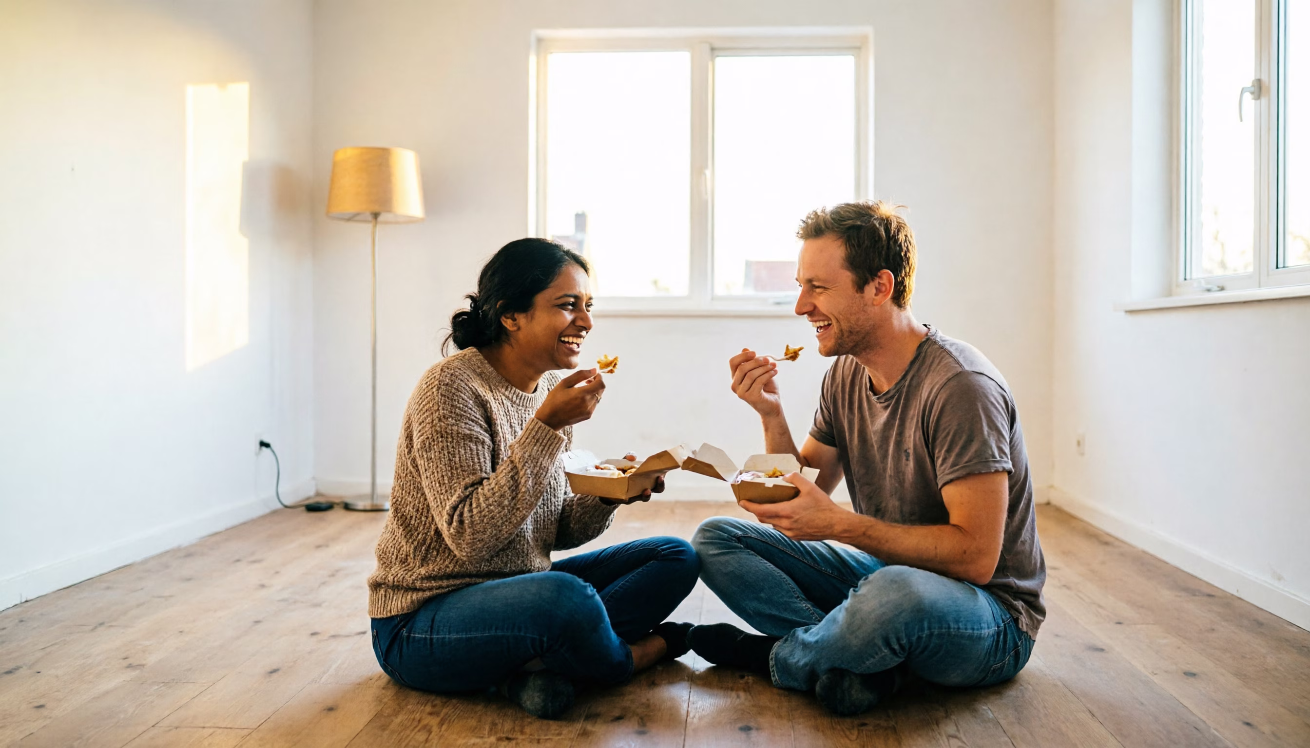 A couple sitting on the floor of the empty living room of their new home, sharing a takeaway meal