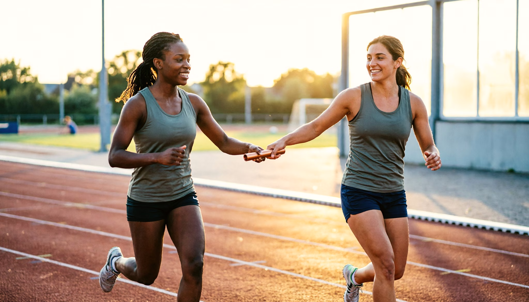 Twee hardloopsters geven het estafettestokje over op de baan van een atletiekclub in avondlicht