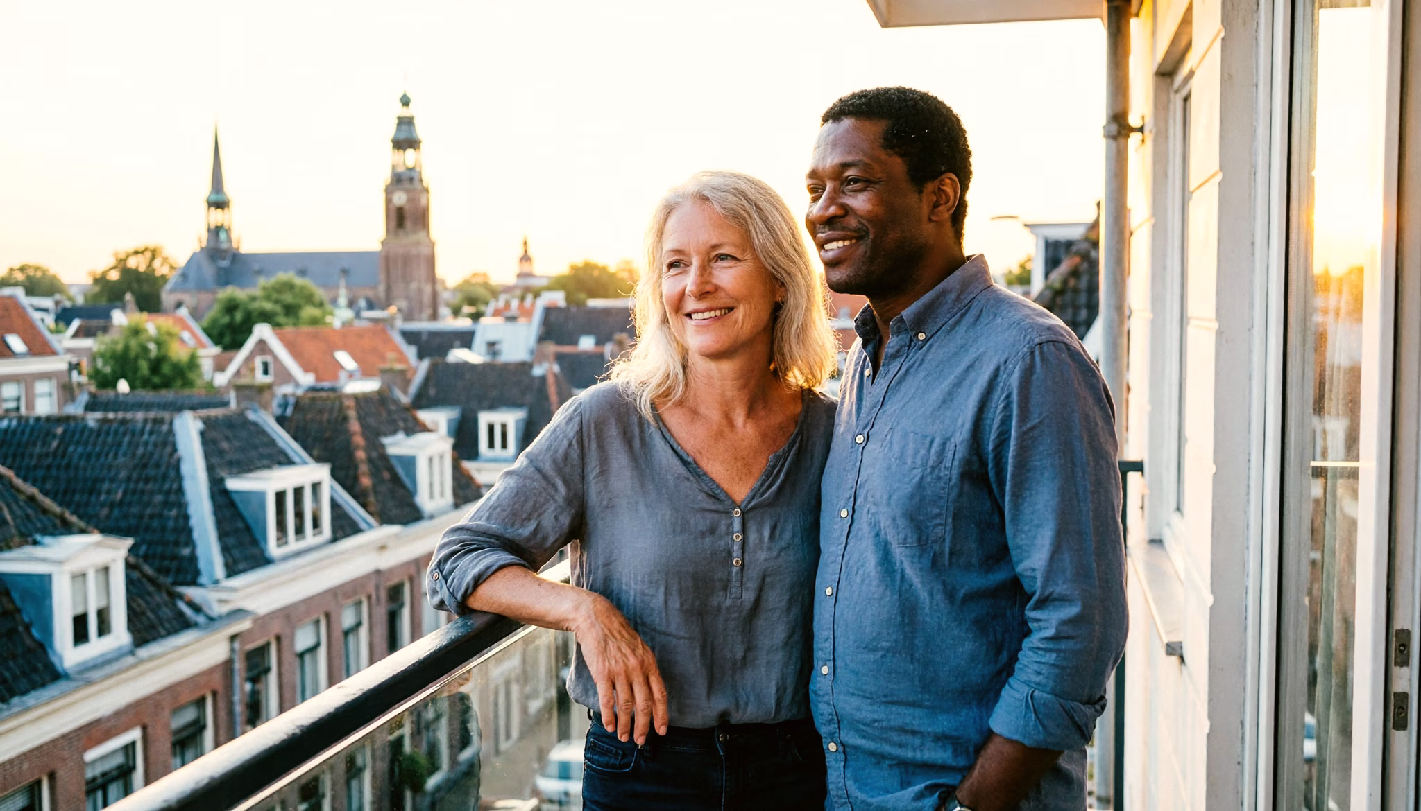 Een stel van midden vijftig staat ontspannen op een balkon met uitzicht over een Nederlandse stad